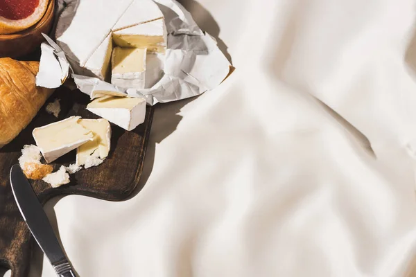 Vista superior del desayuno francés con croissant, Camembert, pomelo sobre tabla de cortar de madera con cuchillo sobre mantel blanco — Stock Photo