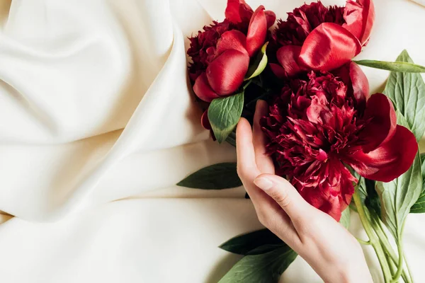 Vista dall'alto di mano femminile e mazzo di peonie rosse su panno bianco — Foto stock