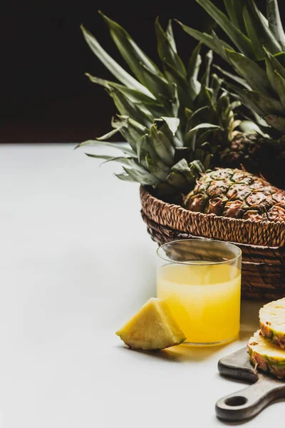 Succo d'ananas fresco in vetro vicino a deliziosi frutti in cesto su superficie bianca isolato su nero — Foto stock