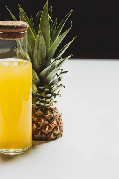 Suco de abacaxi fresco em garrafa perto de cortar deliciosa fruta na superfície branca isolada em preto — Fotografia de Stock