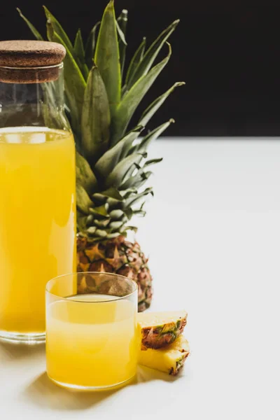 Zumo de piña fresca en botella y vidrio cerca de fruta deliciosa cortada en la superficie blanca aislado en negro - foto de stock