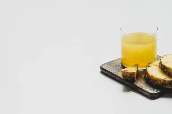 Suco de abacaxi fresco em vidro perto de cortar frutas deliciosas em tábua de corte de madeira no fundo branco — Fotografia de Stock