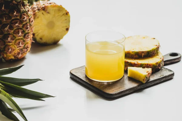 Suco de abacaxi fresco em vidro perto de cortar frutas deliciosas em tábua de corte de madeira no fundo branco — Fotografia de Stock