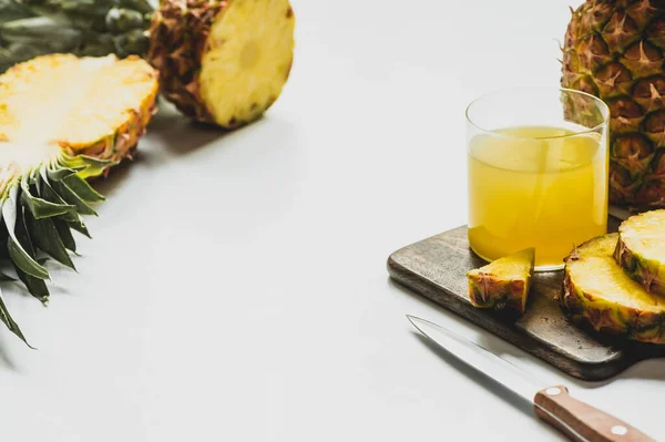 Suco de abacaxi fresco em vidro perto de cortar frutas deliciosas na placa de corte de madeira com faca no fundo branco — Fotografia de Stock