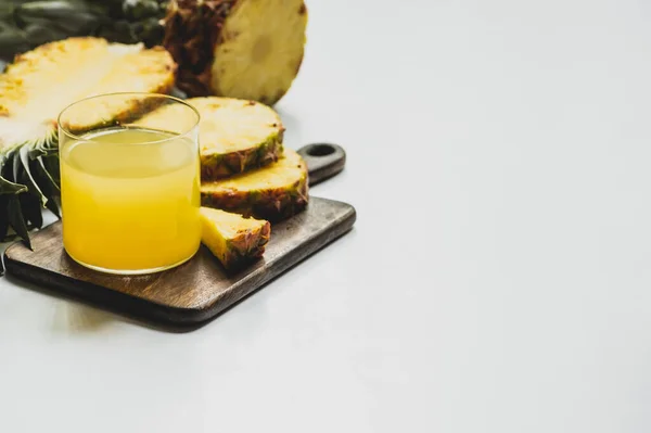 Селективный фокус свежего ананасового сока в стекле рядом с разрезанными вкусными фруктами на деревянной доске для резки на белом фоне — стоковое фото