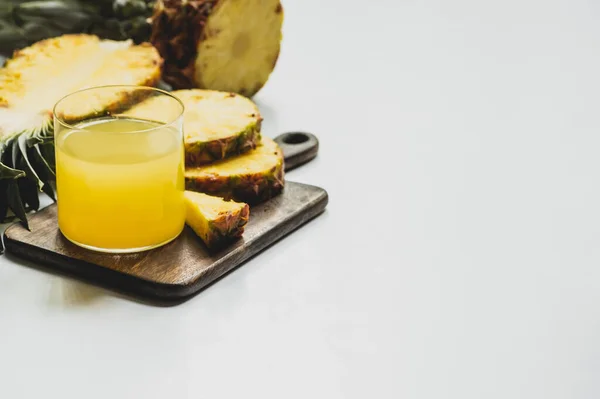 Enfoque selectivo de jugo de piña fresca en vidrio cerca de fruta deliciosa cortada en tabla de cortar de madera sobre fondo blanco - foto de stock