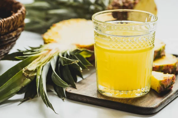 Enfoque selectivo de jugo de piña fresca en vidrio cerca de fruta deliciosa cortada en tabla de cortar de madera sobre fondo blanco - foto de stock