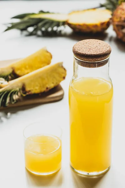 Foco seletivo de suco de abacaxi fresco em garrafa e vidro perto de cortar frutas deliciosas na placa de corte no fundo branco — Fotografia de Stock