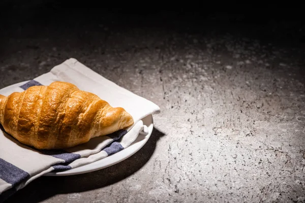 Croissant cozido no forno fresco na toalha e placa na superfície cinzenta concreta no escuro — Fotografia de Stock