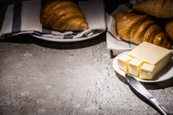 Selective focus of fresh baked croissants on towel near butter and knife on concrete grey surface in dark — Stock Photo