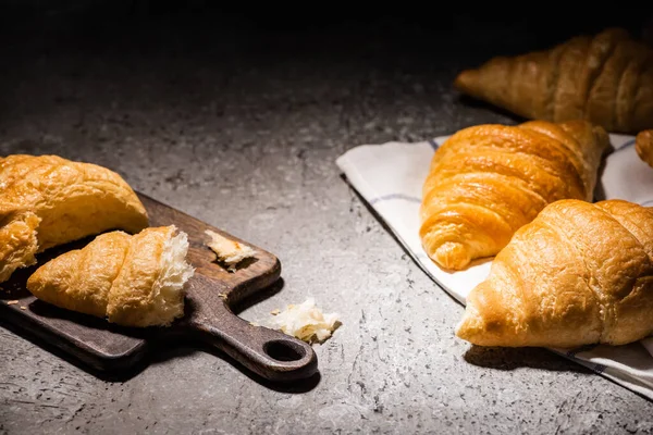 Croissants cozidos no forno fresco na toalha perto da placa de corte na superfície cinzenta concreta no escuro — Fotografia de Stock