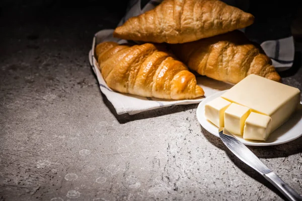 Croissant appena sfornati su asciugamano vicino al burro e coltello su una superficie grigio cemento al buio — Foto stock