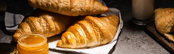 Fresh baked croissants on towel near jam on concrete grey surface in dark, panoramic shot — Stock Photo