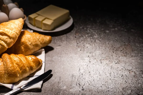 Foco seletivo de croissants frescos assados com faca na toalha perto da manteiga e ovos na superfície cinza de concreto no escuro — Fotografia de Stock