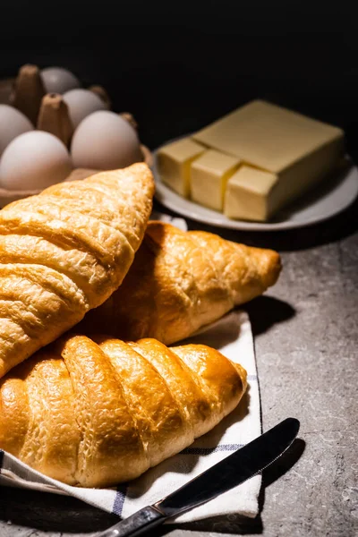 Enfoque selectivo de croissants recién horneados con cuchillo en la toalla cerca de la mantequilla y huevos en la superficie gris de hormigón en la oscuridad - foto de stock