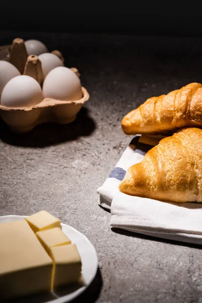 Selective focus of fresh baked croissants on towel near butter and eggs on concrete grey surface in dark — Stock Photo