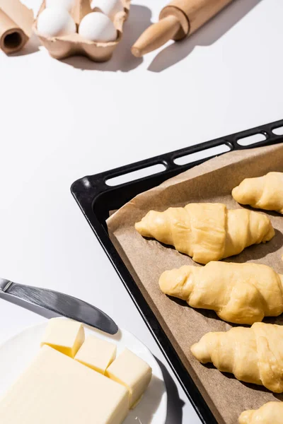 Selective focus of raw croissants on baking tray near rolling pin, parchment paper, butter, eggs, knife on white background — Stock Photo