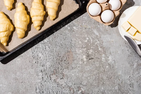 Vue de dessus des croissants non cuits sur la plaque à pâtisserie près des œufs et du beurre sur la surface gris béton — Photo de stock