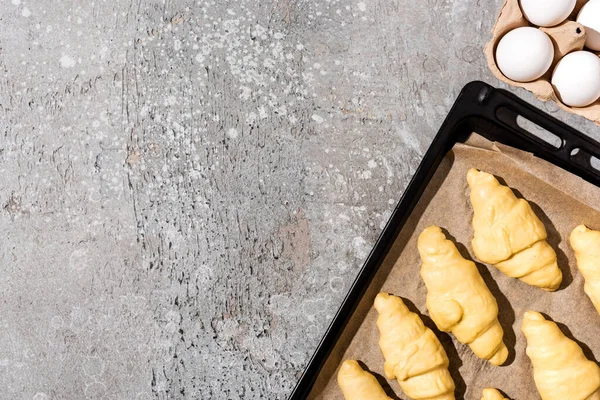 Vista superior de cruasanes sin cocer en bandeja para hornear cerca de los huevos en la superficie gris de hormigón — Stock Photo