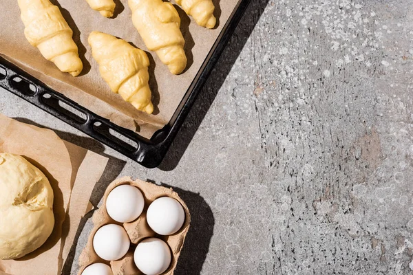 Vista superior de croissants não cozidos na assadeira perto de ovos e massa na superfície cinza de concreto — Fotografia de Stock