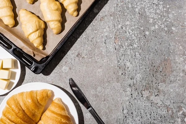 Vista superior de croissants não cozidos na bandeja de cozimento perto cozido na placa e manteiga na superfície cinzenta concreta — Fotografia de Stock
