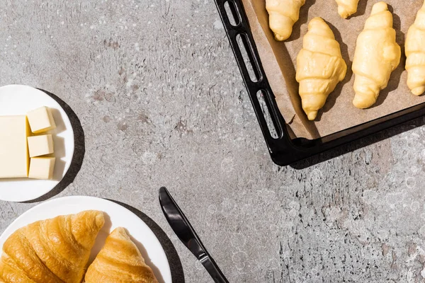 Vista dall'alto di croissant crudi su teglia vicino cotto su piatto e burro su superficie grigio cemento — Foto stock
