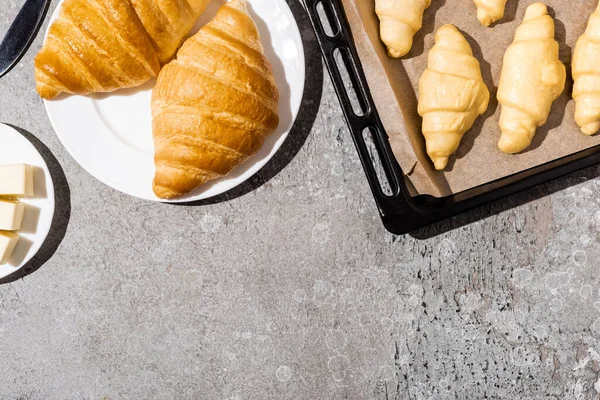 Vista superior de croissants sin cocer en bandeja para hornear cerca de cocinado en plato y mantequilla en superficie gris hormigón - foto de stock