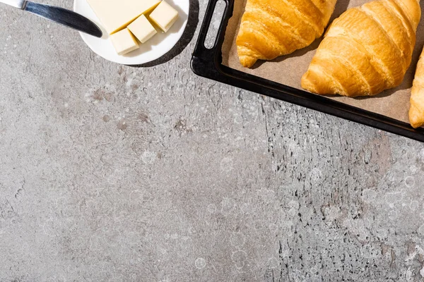 Vista superior de croissants deliciosos horneados en bandeja para hornear cerca de la mantequilla en la superficie gris de hormigón - foto de stock