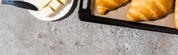 Vue de dessus de délicieux croissants cuits sur plaque à pâtisserie près du beurre sur une surface gris béton, vue panoramique — Photo de stock