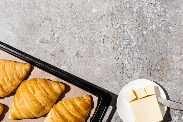 Vue de dessus de délicieux croissants cuits sur plaque à pâtisserie près du beurre sur une surface gris béton — Photo de stock