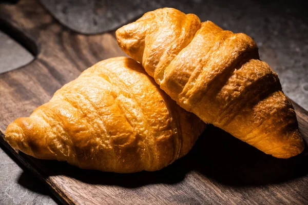 Fresh baked croissants on wooden cutting board on concrete grey surface in dark — Stock Photo