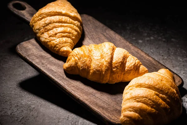 Fresh baked croissants on wooden cutting board on concrete grey surface in dark — Stock Photo
