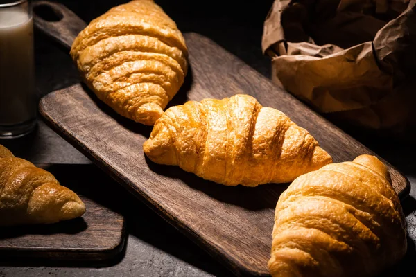 Foco seletivo de croissants cozidos no forno fresco na toalha e na placa de corte de madeira perto do saco de papel e do leite na superfície cinzenta concreta no escuro — Fotografia de Stock