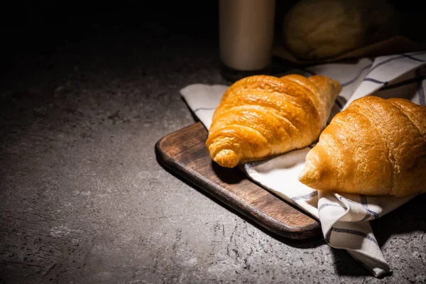 Fresh baked croissants on towel and wooden cutting board on concrete grey surface in dark — Stock Photo