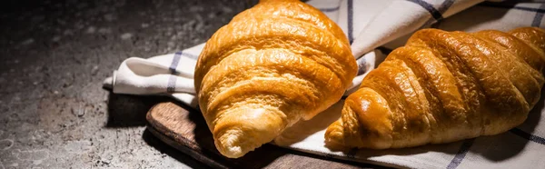 Croissants cozidos no forno fresco na toalha e na placa de corte de madeira na superfície cinzenta concreta no tiro escuro, panorâmico — Fotografia de Stock