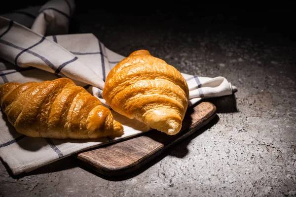 Croissant appena sfornati su asciugamano e tagliere in legno su superficie grigio cemento scuro — Foto stock