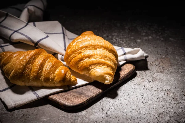 Croissants cozidos no forno fresco na toalha e na placa de corte de madeira na superfície cinzenta concreta no escuro — Fotografia de Stock