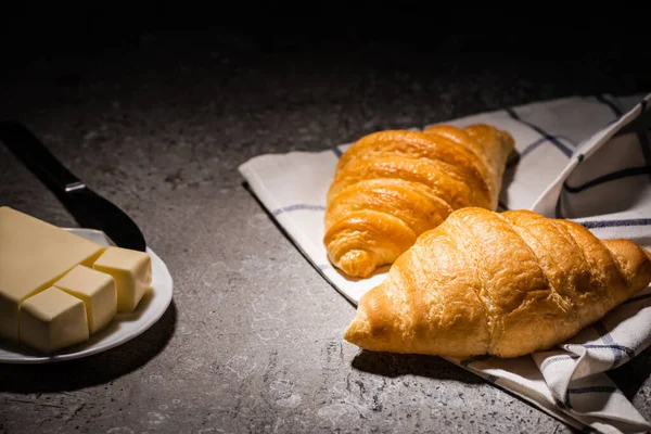 Frisch gebackene Croissants auf Handtuch in Butternähe und Messer auf betongrauer Oberfläche in Dunkelheit — Stockfoto