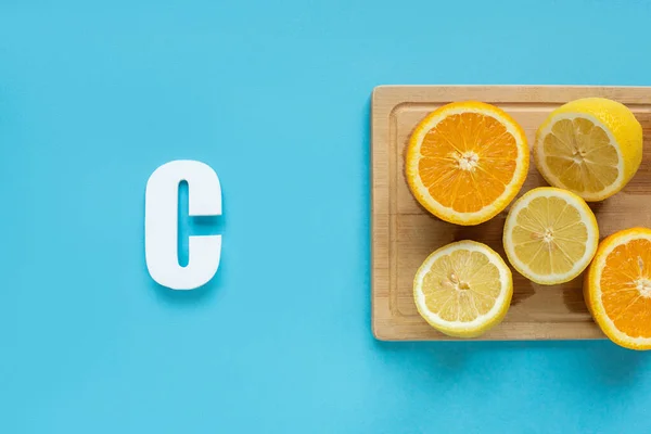 Top view of ripe cut lemon and orange on wooden cutting board near letter C on blue background — Stock Photo