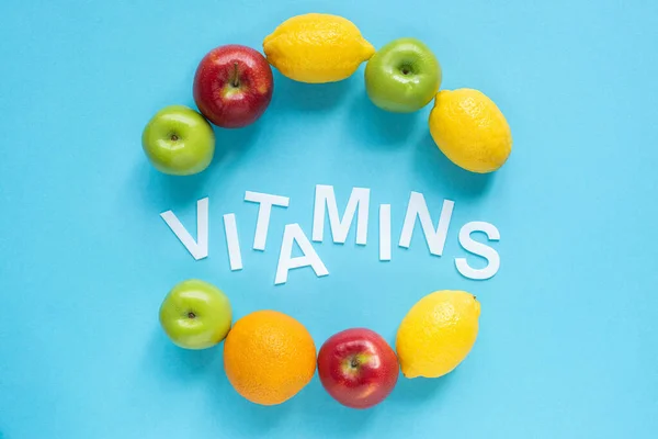 Vista superior de frutas maduras alrededor de las vitaminas de la palabra sobre fondo azul - foto de stock