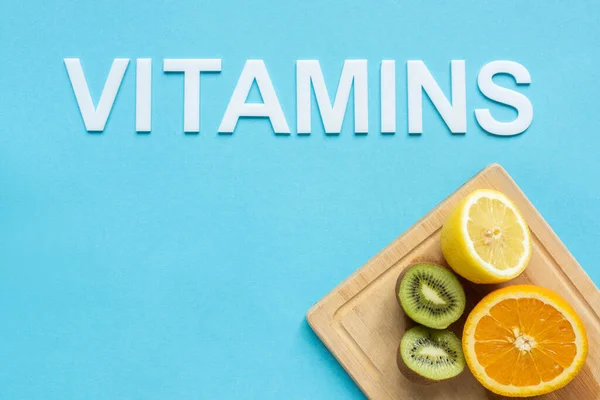 Top view of ripe orange, lemon and kiwi halves on wooden cutting board and word vitamins on blue background — Stock Photo