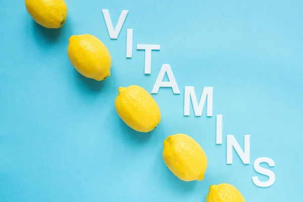 Flat lay com limões amarelos maduros e vitaminas palavra no fundo azul — Fotografia de Stock