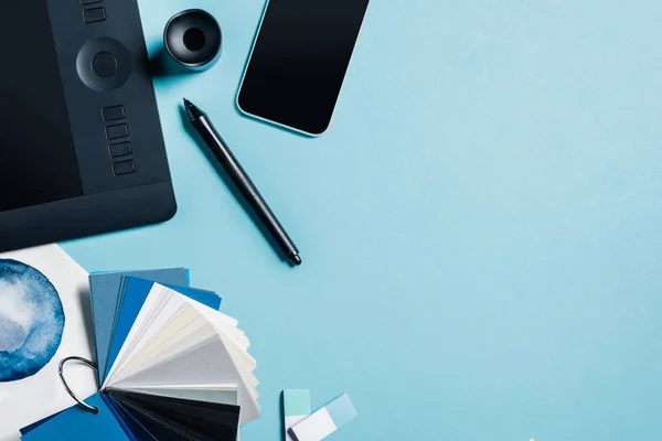 Вид сверху графического планшета, смартфона, цветных образцов и акварельного рисунка на синем фоне — стоковое фото