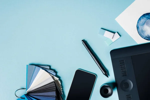 Vue de dessus des échantillons de couleur, smartphone près de la tablette graphique et dessin à l'aquarelle sur la surface bleue — Photo de stock