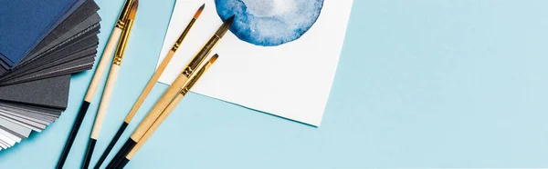 Recadrage panoramique de pinceaux près de l'aquarelle dessin et des échantillons de couleur sur la surface bleue — Photo de stock