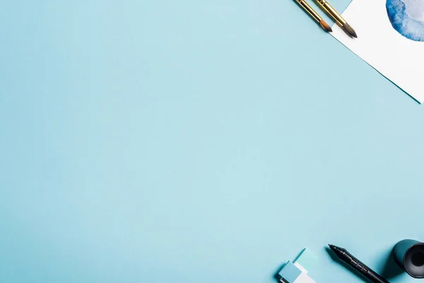 Vue du dessus des pinceaux près du dessin à l'aquarelle et du stylet de la tablette graphique sur fond bleu — Photo de stock