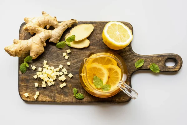 Vista superior del té caliente cerca de raíz de jengibre, limón y menta sobre tabla de cortar de madera sobre fondo blanco - foto de stock