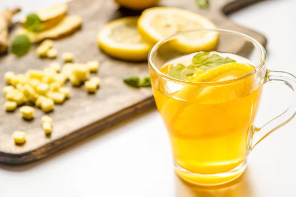 Foyer sélectif de thé chaud dans le verre près de la racine de gingembre, citron et menthe sur planche à découper en bois sur fond blanc — Photo de stock