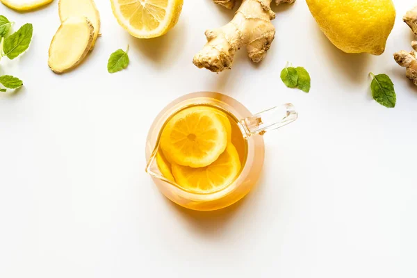 Vue du dessus du thé chaud dans une théière en verre près de la racine de gingembre, citron et menthe sur fond blanc — Photo de stock