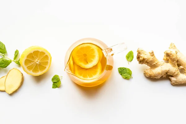 Vista superior de té caliente en tetera de vidrio cerca de raíz de jengibre, limón y menta sobre fondo blanco - foto de stock